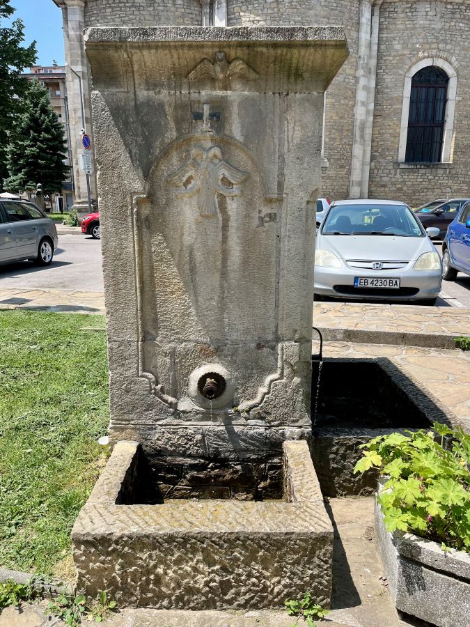 Каменна градска чешма с два чучура, издялан надпис „1762“ и кръстове върху каменния фронтон, заснета на калдъръмен площад в старинен градски център.