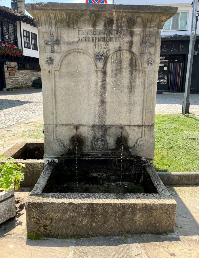 Каменна градска чешма с два чучура, издялан надпис „1762“ и кръстове върху каменния фронтон, заснета на калдъръмен площад в старинен градски център.
