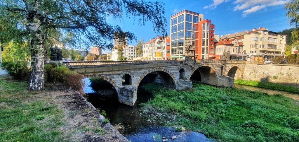 Снимка на Баев мост в Габрово – стар каменен арков мост над реката в централната градска част, с сгради и синьо небе на заден план.