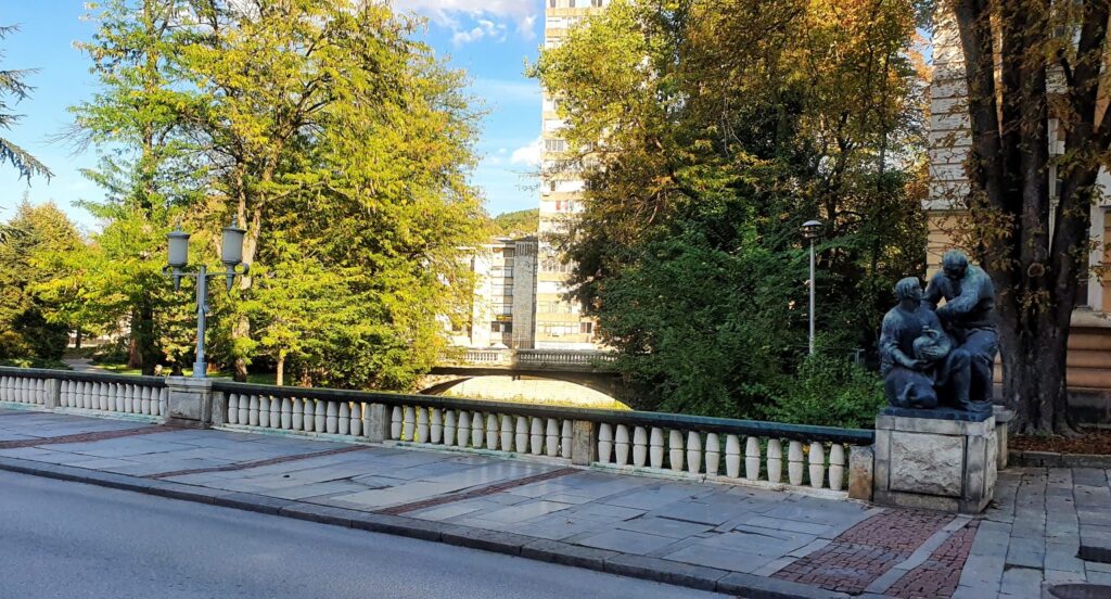 Снимка на Грънчарския мост в Габрово – градски мост с балюстрада и скулптурни фигури край реката, заобиколен от дървета и жилищни сгради.