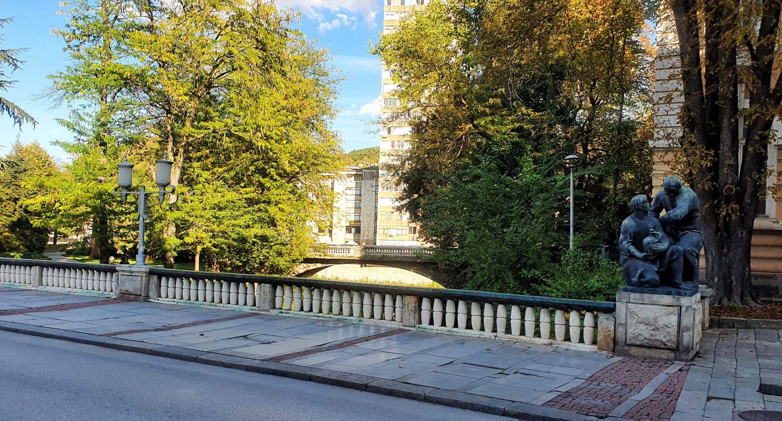 Снимка на Грънчарския мост в Габрово – градски мост с балюстрада и скулптурни фигури край реката, заобиколен от дървета и жилищни сгради.