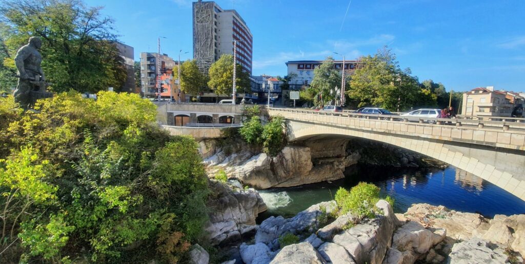 Снимка на мост „Игото“ в Габрово – каменен мост над река Янтра при скалите Грамадата, с градски сгради и зеленина наоколо.