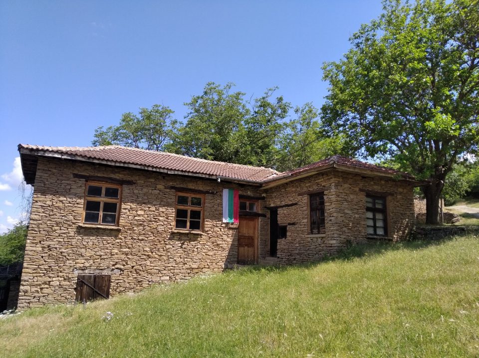 Каменна обществена сграда с българско знаме в село Иглика, заснета в слънчев летен ден.