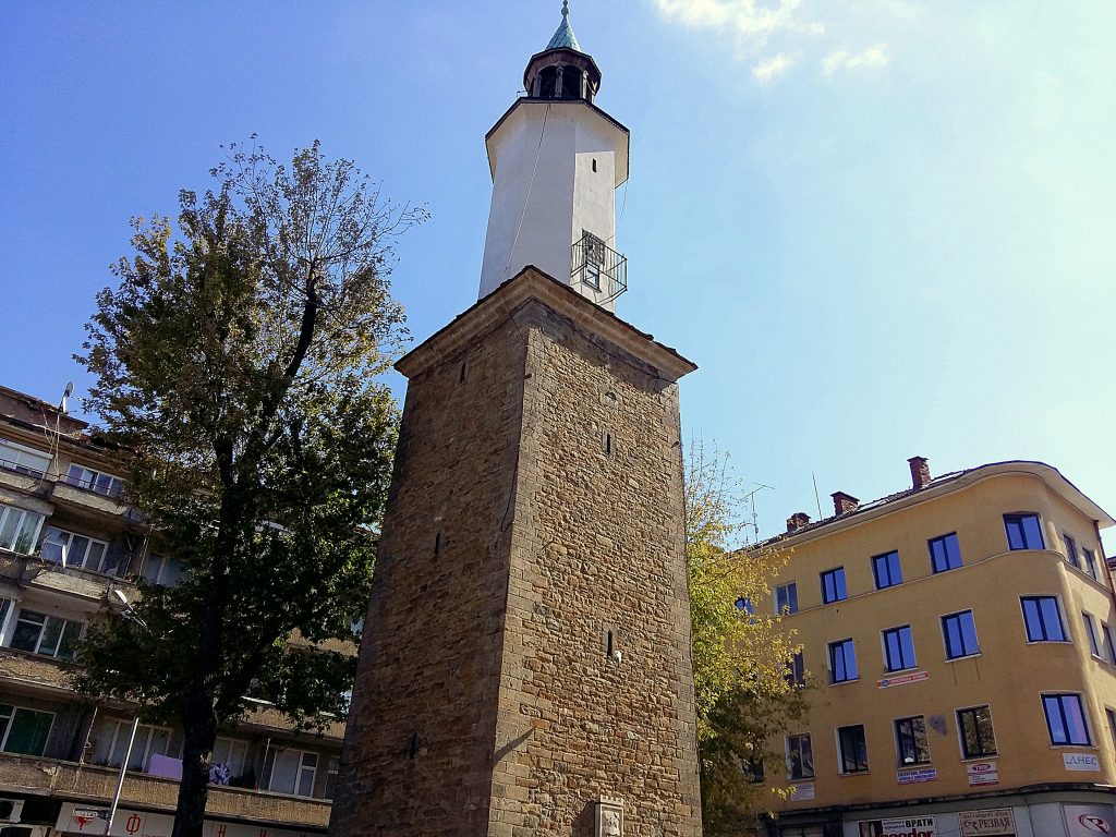 Снимка на Габровската часовникова кула в центъра на Габрово, България – каменна кула с бяла надстройка и градски сгради наоколо.