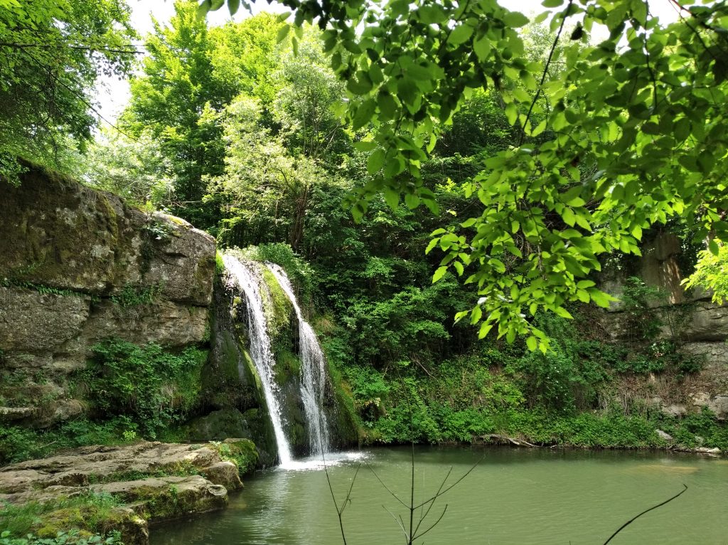 Иванилският водопад пада между скали в зелена горска местност, с тих вир в подножието.