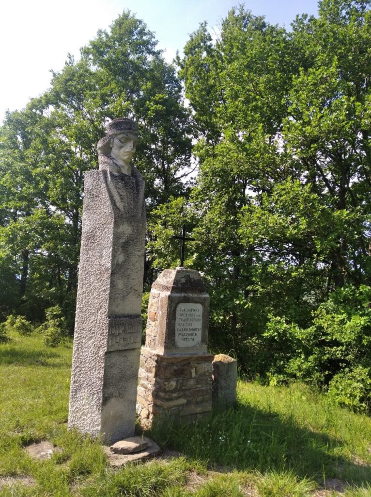 Каменен паметник с кръст сред зелена горска местност.